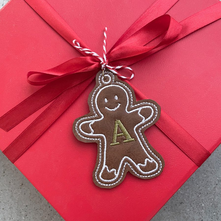 In-the-hoop machine embroidery design of a gingerbread cookie gift tag, featuring a personalized letter "A" initial, with intricate stitching and festive details.