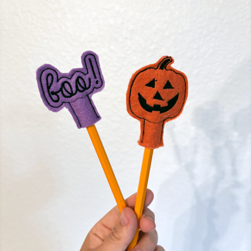 Person holding two Halloween embroidery pencil covers made with felt and thread – featuring a pumpkin and 'BOO' design.